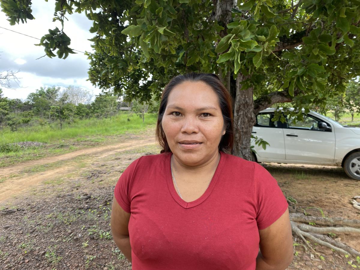 Francilene dos Santos Pereira, da AMYK, posa para foto. Ela usa uma camiseta vermelha e atrás dela tem uma árvore e um carro