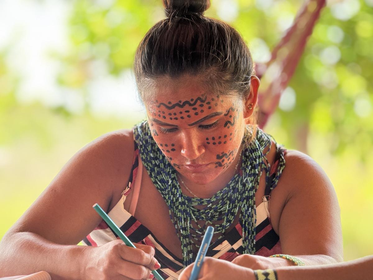 Ariane Yanomami aparece sentada, escrevendo em um papel. Ela tem o rosto pintado