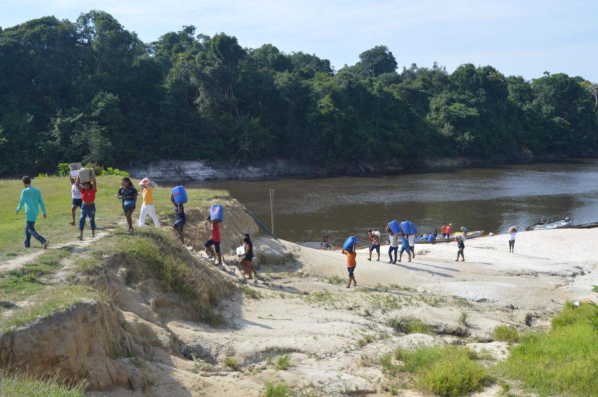 Indígenas saem de rio carregando baldes com água. Ao fundo, há uma floresta