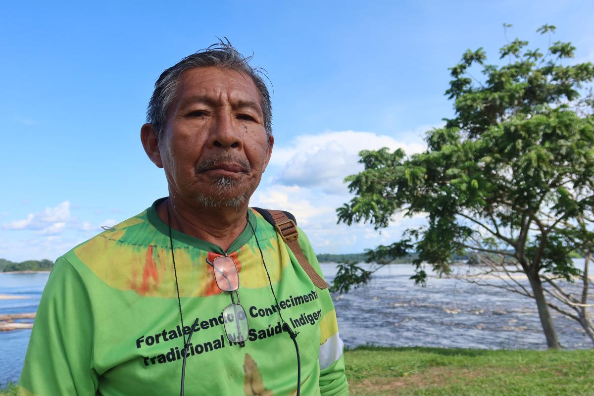 No Alto Rio Negro, Arlindo Maia Tukano olha para a câmera. Atrás dele, um rio e uma árvore do lado direito
