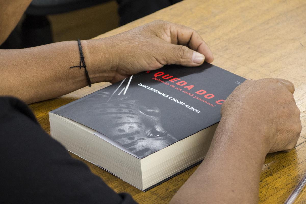 Imagem mostra mãos de Davi Kopenawa segurando seu livro "A Queda do Céu"