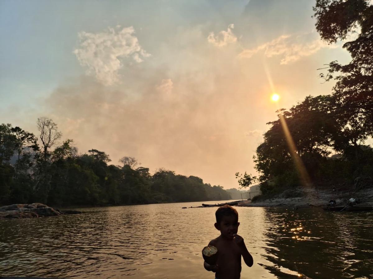 Imagem mostra uma criança na frente de um rio. Atrás dela e do rio, uma fumaça é vista